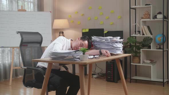 Tired Asian Man Sleeping Due To Working Hard With Documents At The Office alt