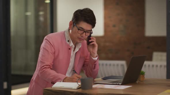 Smartly dressed asian businessman sitting at desk using laptop taking notes alt