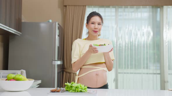 Portrait of Asian young pregnant woman hold salad in bowl in kitchen. alt