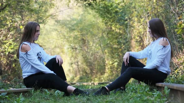 Twin Girls Sit on Train Tracks Opposite Each Other and Look at Camera in Nature alt