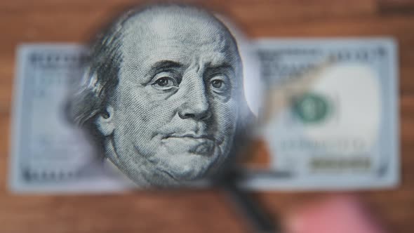 One Hundred Dollars Inspects Under Magnifying Glass on Wooden Table alt