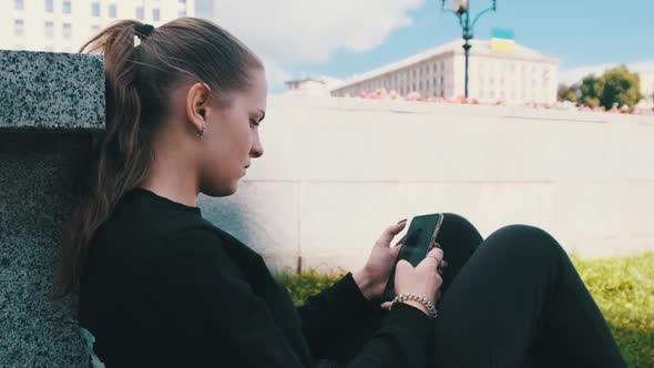 Young Woman Uses Smartphone While Sitting on the Parapet in the Street Park alt