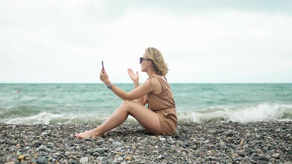 Girl Talking on Video Call Sitting on the Beach on the Background of the Ocean with Waves