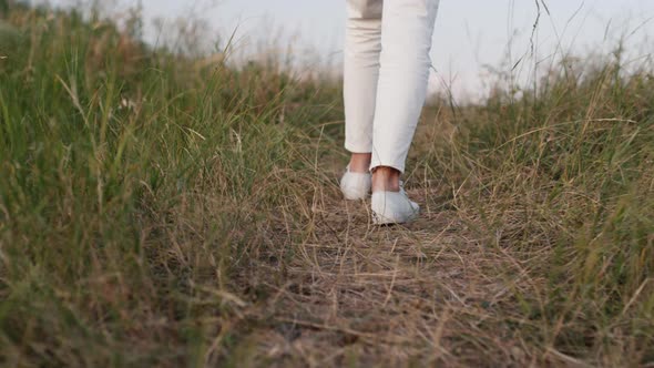 The Legs of a Woman Dressed in White Clothes Walk Along the Path Among the Grass alt