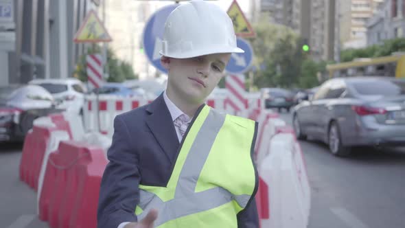 Tired Little Boy in Constructor Helmet on His Head, and Uniform Looking in Camera Showing Thumb Up alt