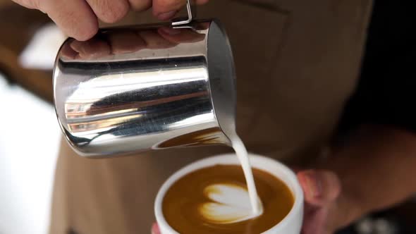 Close up shot of professional barista pouring milk from pitcher into coffee cup making latte art