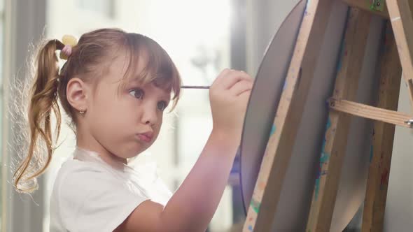 Beautiful Concentrated Little Girl Paints with Paintbrush on Canvas on an Easel