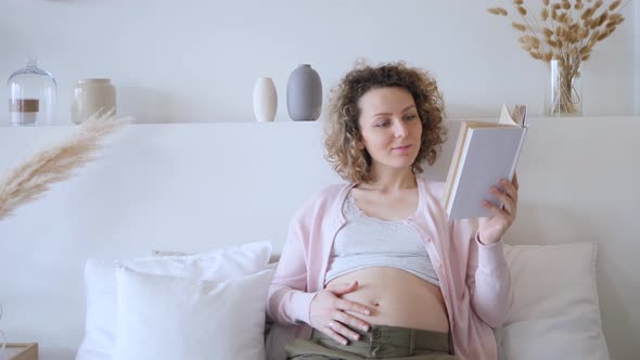 Smiling Pregnant Girl Reading Book In Bed At Home In Bedroom alt