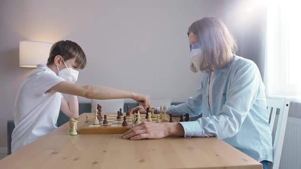 Father and Son in Medical Mask on Selfisolation They Play Chess Sitting in the Living Room alt