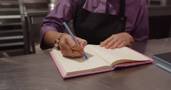 A confectioner girl draws sketches of a cake in a notebook while sitting in a pastry shop