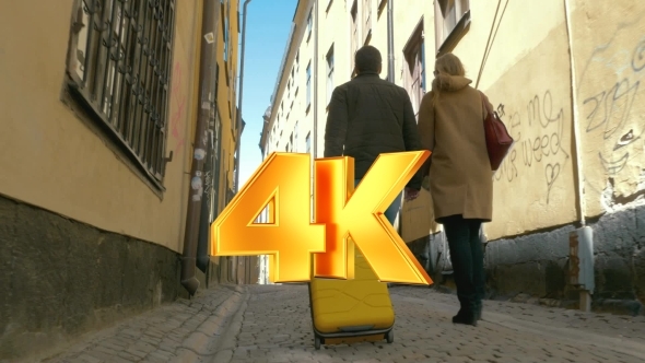 Couple Of Tourists With Roll-on Bag Walking In Old alt