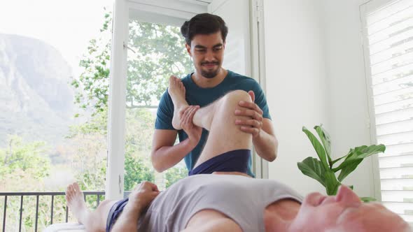 Biracial young male therapist treating leg of retired senior man at health club alt