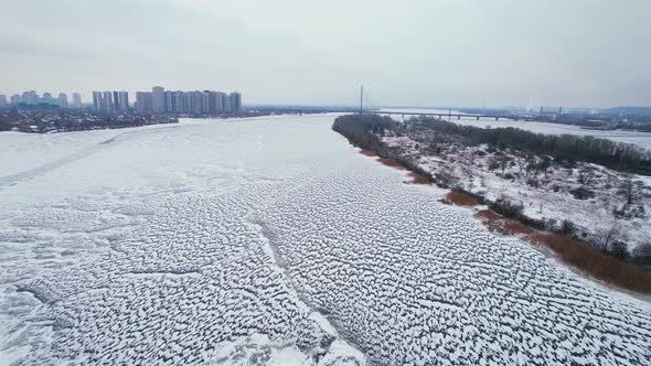 Aerial View of a Frozen River with a Bridge and City Buildings on the Shore