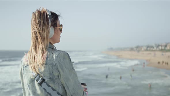 Stylish Happy Young Woman in Denim Jacket and Headphones Looking at Ocean Beach alt