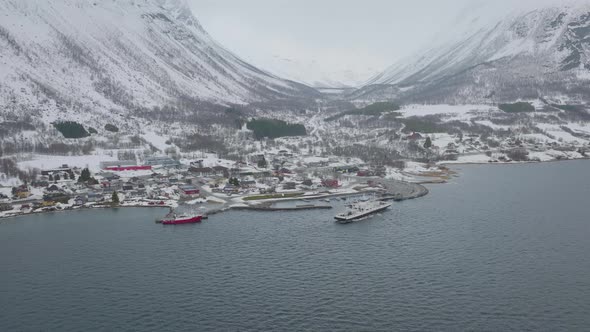 Ferryboat departs Olderdalen town at foot of foggy, imposing mountains; aerial alt