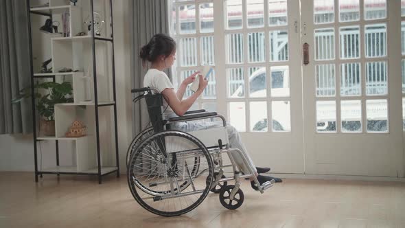 Asian Woman Sitting In A Wheelchair Reading Book Near Door In The House alt