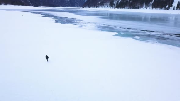 The drone orbits around a young east european guy with a camera who walks through a snowy field. In alt