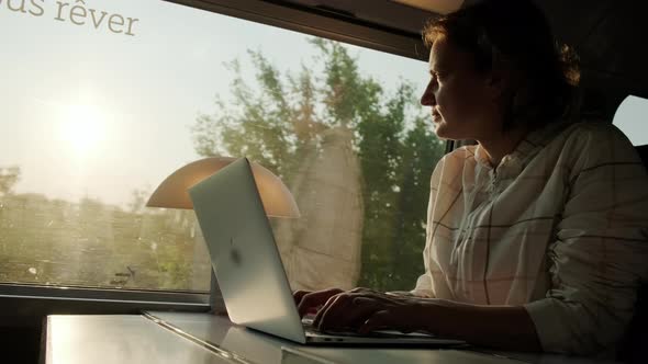 Woman Works on Laptop While Riding Train Sitting Near Window on Sunny Day alt
