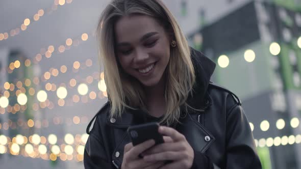 Young Girl Uses a Smartphone Sitting Outside in a Modern Residential Area Evening in the City Blurry alt