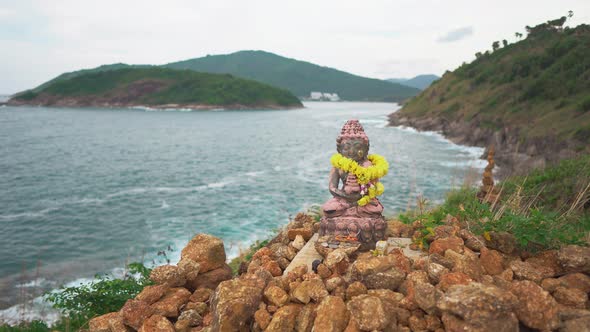 Little Statue of Sitting in A Lotus Position Deity on Top of A Cliff Against the Sea and Green Hills alt