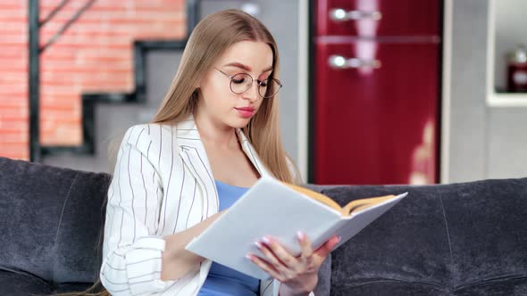 Focused Young Beautiful Girl in Glasses Reading Paper Book Relaxing on Sofa Medium Shot alt