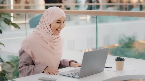Happy Young Arab Girl in Hijab Sitting at Table Answering Video Call on Laptop Joyfully Waving Hand alt