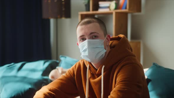 Portrait of Young American Man Sitting on Sofa Wearing Medical Surgical Mask to Protect Coronavirus alt