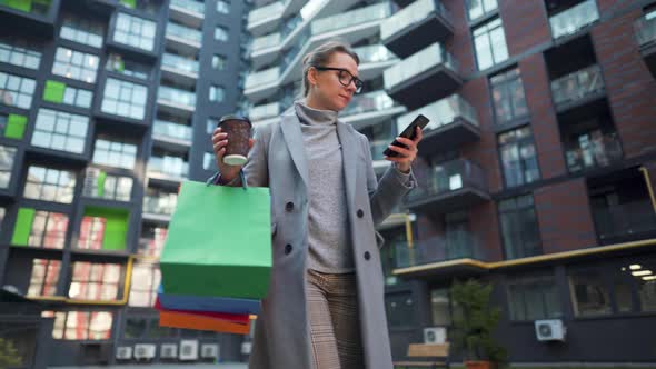 Woman Walking on a Business District Holds Takeaway Coffee Shopping Bags and Using Smartphone alt
