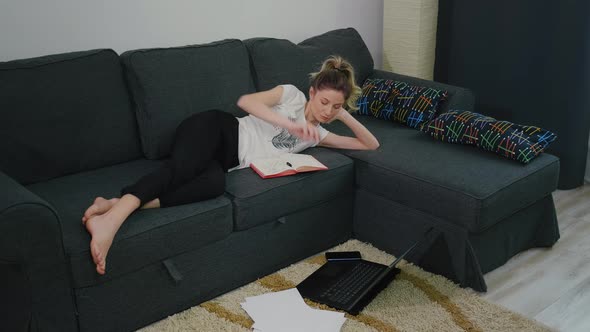 Pretty woman makes notes in notebook while lying on couch at home in a modern apartment alt