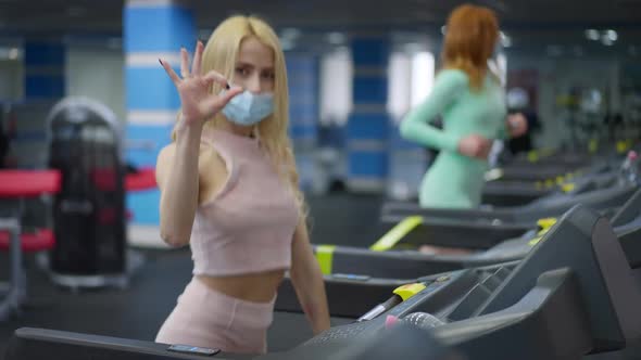 Joyful Confident Sportswoman in Coronavirus Face Mask Showing OK Sign Looking at Camera and Running alt