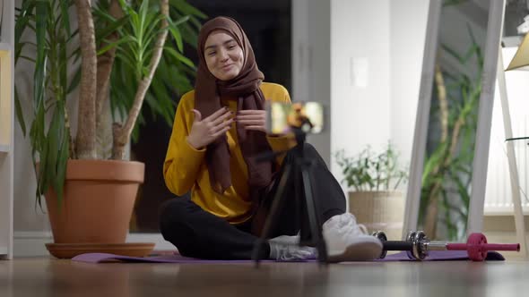 Cheerful Sport Blogger in Hijab Waving and Talking at Smartphone Streaming Online alt