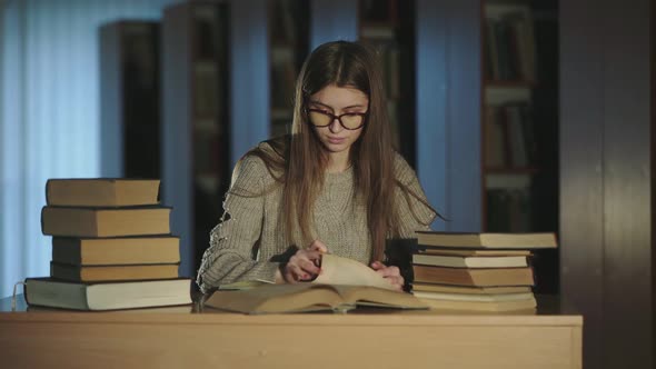 Pretty Girl in Glasses Reads a Book at Desk in Library, Crosses Hands and Smiles alt