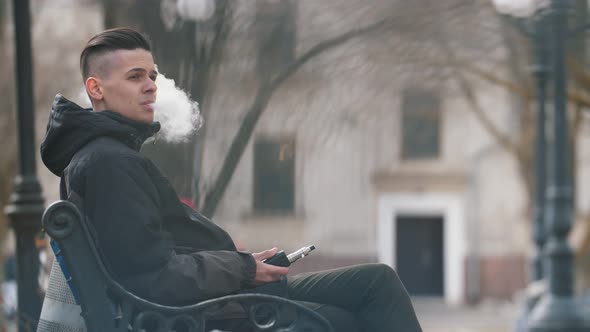 Snappy Brunet Man Sitting and Smoking E-cigarette Outdoors in a Park in Spring 