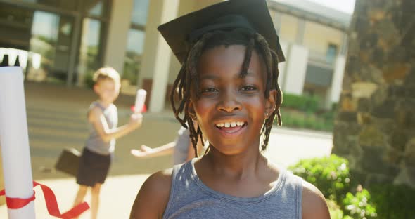 Video of happy african american boy wearing graduation hat and holding diploma in front of school alt