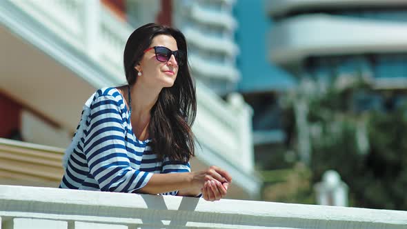 Happy Smiling Female Tourist in Sunglasses and Striped Dress Putting Hands on White Railing alt