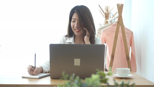 young smiling asian woman working on laptop while sitting in a living room at home using phone. alt