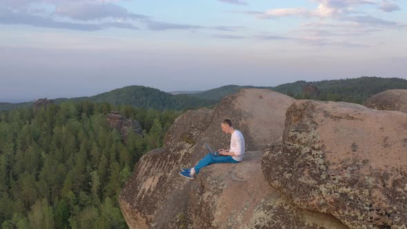 A Man Is Typing on a Laptop Sitting on the Edge of a Rocky Mountain.