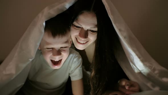 Young Woman Mom and Her Son Watching a Funny Film Together on Tablet and Laugh Under the Blanket.