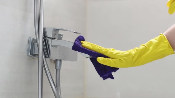 The hands of a woman in yellow gloves are wiping a wet faucet with a lilac rag.