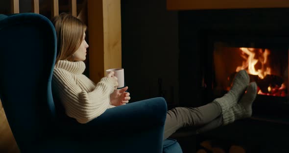 Relaxed Woman Warms at Fireplace Sitting in Armchair and Drinking Hot Tea