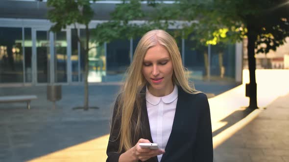 Cheerful Business Woman with Phone Outdoors. Content Blond Lass in Black Jacket Holding Smartphone alt