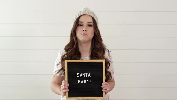 Woman frowning then smiling as she holds sign saying, Santa Baby alt