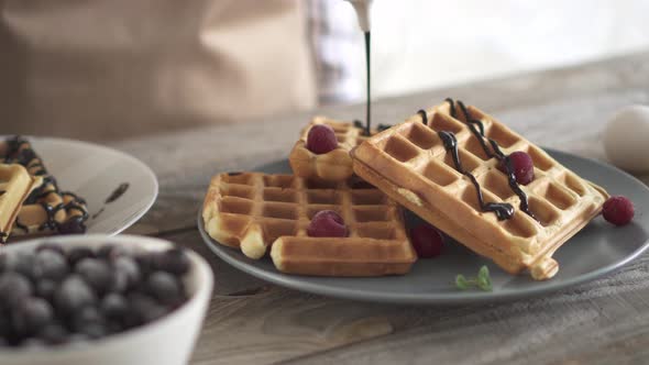 Two Plates Of Waffles. A Girl Pours Chocolate Sauce Over Belgian Waffles. Sweet Belgian Waffles  alt