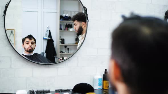 A Man in a Chair in a Hairdressing Salon Looks at Himself in the Mirror While the Hairdresser Combs