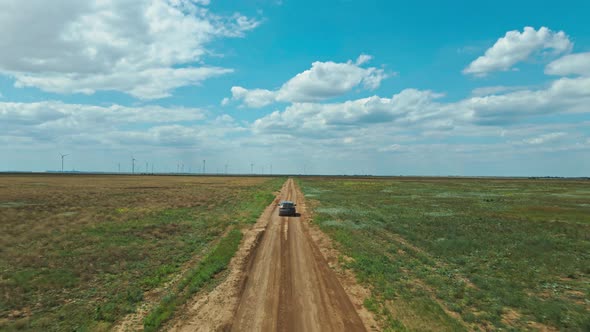 Drone Following Car Moving By Rustic Road Through Green Fields on Background of Wind Turbines alt