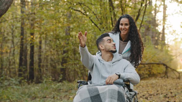 Smiling Nurse Girl in Medical Gown Pushing Young Guy in Wheelchair Walking in Hospital Park Enjoy alt