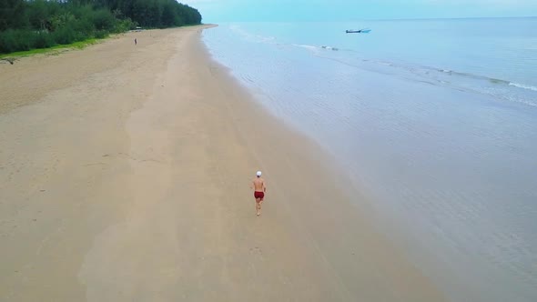 Drone view of a man running by a perfect beach with a boat in the background alt