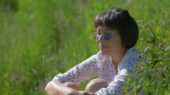 Woman in Colorful Sunglasses Enjoys Sunlight and Flower Fragrance on Grass Field alt
