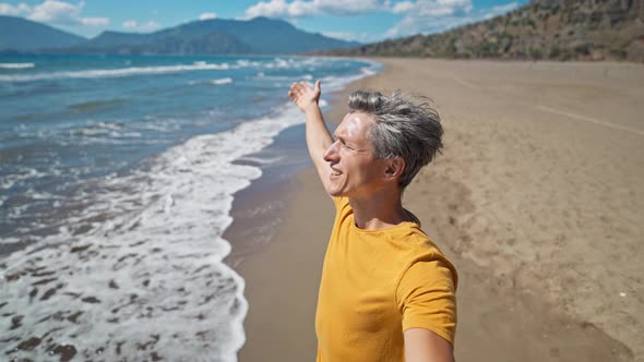 Happy Middle Aged Greyhead Man Stands at Sandy Sea Coast with Raised Hands alt