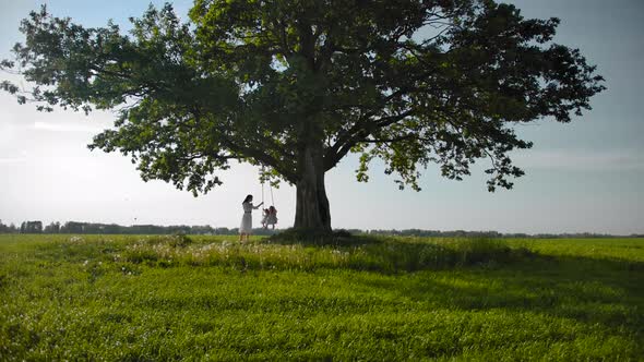 Lonely Beautiful Big Tree and Tiny Silhouettes of Mom with Two Daughters Swinging on a Swing alt